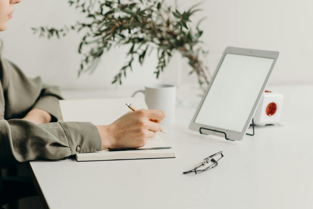 Freelancer writing notes in a minimalist home office with a white tablet, plant, and reading glasses.